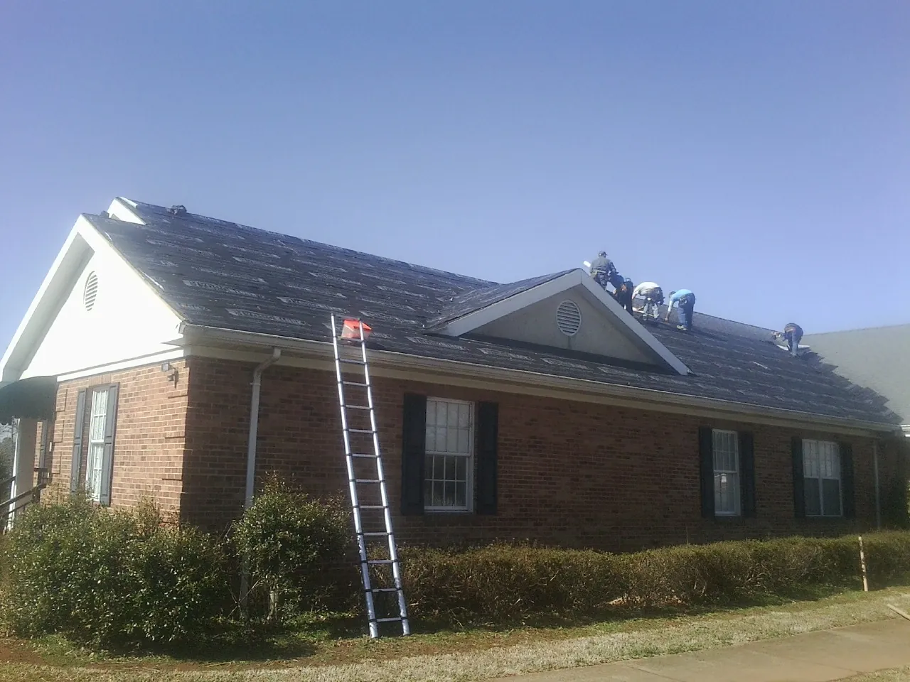 Crew tearing shingles off a brick ranch roof with a ladder set — Emergency Roof Repair work by our crew in Spring Grove